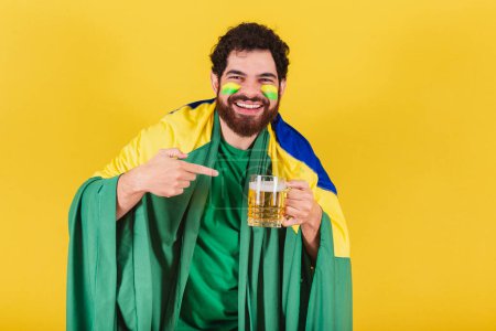 Photo for Caucasian man with beard, brazilian, soccer fan from brazil, holding and pointing to glass of beer. - Royalty Free Image