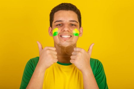 Foto de Joven aficionado al fútbol brasileño. vestido de verde, y amarillo, pulgares hacia arriba, como - Imagen libre de derechos