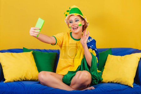 Foto de Mujer caucásica, pelirroja, aficionado al fútbol brasileño, brasileño, en el sofá en la videollamada smartphone - Imagen libre de derechos