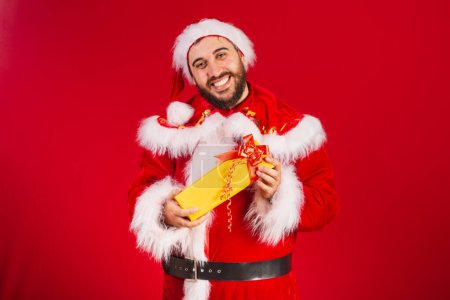 Photo for Brazilian man, dressed in santa claus clothes, giving yellow gift to camera. - Royalty Free Image