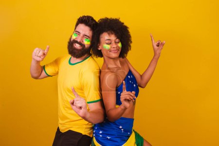Foto de Pareja de aficionados brasileños al fútbol, vestidos con los colores de Brasil, mujer negra, hombre caucásico. bailar, divertirse. - Imagen libre de derechos