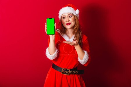 Foto de Hermosa brasileña pelirroja, vestida con ropa de Navidad, Santa Claus. mostrando smartphone con pantalla croma verde. - Imagen libre de derechos