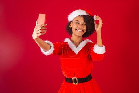Téléchargez les photos : Belle femme brésilienne noire, habillée en Père Noël claus, maman claus, prenant autoportrait avec smartphone - en image libre de droit