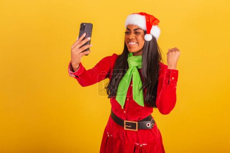 Foto de Hermosa mujer brasileña negra, vestida con ropa de Navidad, Santa Claus, smartphone y celebrando. - Imagen libre de derechos