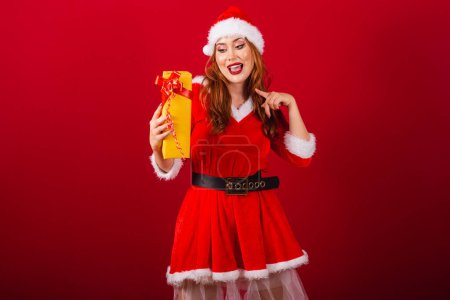 Photo for Beautiful Brazilian red-haired woman, dressed in Christmas clothes, Santa Claus. showing and pointing to yellow present. - Royalty Free Image