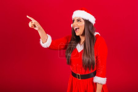 Foto de Hermosa mujer brasileña vestida con ropa de Navidad, Santa Claus. apuntando a algo en la distancia. - Imagen libre de derechos