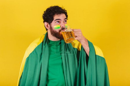Foto de Hombre caucásico con barba, brasileño, fanático del fútbol brasileño, bebiendo cerveza de la taza. buen concepto de cerveza. - Imagen libre de derechos