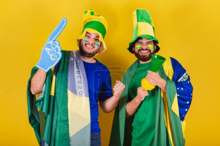 Foto de Dos amigos, brasileños, aficionados al fútbol de Brasil, utilizando la bandera de Brasil, cuerno y dedo para animar en el partido de fútbol. - Imagen libre de derechos