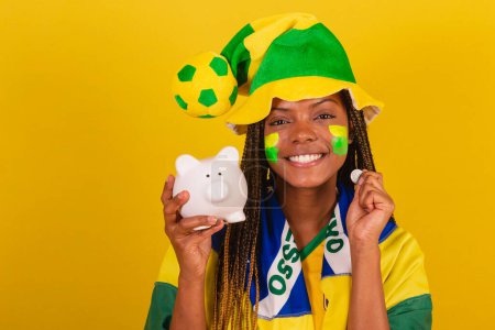 Photo for Black woman young brazilian soccer fan. holding piggy bank and coin. concept of finance, economy. - Royalty Free Image