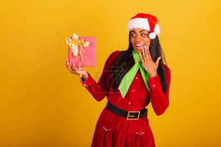 Téléchargez les photos : Belle femme brésilienne noire, vêtue de vêtements de Noël, Père Noël claus, doute avec un cadeau rouge. - en image libre de droit