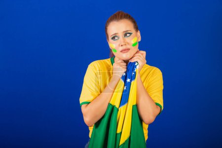 Téléchargez les photos : Femme caucasienne, rousse, fan de football brésilien, brésilien, fond bleu, triste et contrarié avec le drapeau dans les mains - en image libre de droit
