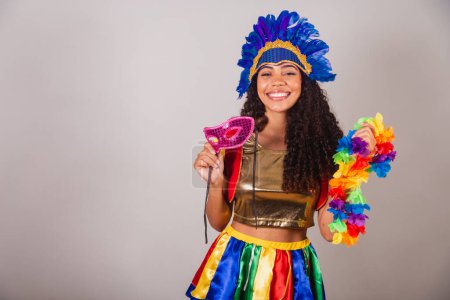 Foto de Hermosa mujer brasileña negra, con ropa frevo, carnaval. en tocado de plumas de carnaval. con máscara de carnaval y collar en las manos. - Imagen libre de derechos