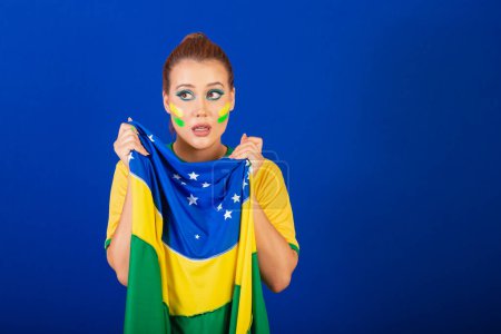 Téléchargez les photos : Femme caucasienne, rousse, fan de football brésilien, brésilien, fond bleu, anxieux et affligé avec le drapeau dans les mains - en image libre de droit