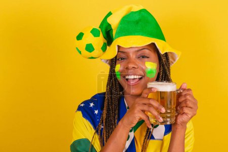 Foto de Mujer negra joven aficionada al fútbol brasileño. beber cerveza y celebrar. - Imagen libre de derechos