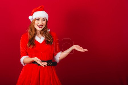 Photo for Beautiful Brazilian red-haired woman, dressed in Christmas clothes, Santa Claus. showing something to the right. - Royalty Free Image