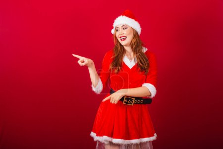 Téléchargez les photos : Belle femme rousse brésilienne, vêtue de vêtements de Noël, le Père Noël. montrer quelque chose à gauche. - en image libre de droit