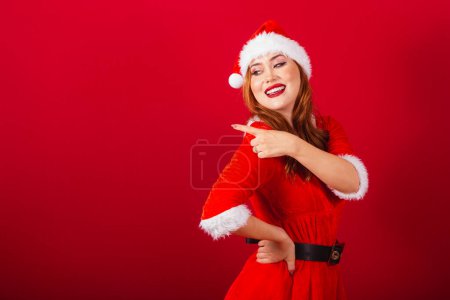 Foto de Hermosa brasileña pelirroja, vestida con ropa de Navidad, Santa Claus. mostrando algo a la izquierda. - Imagen libre de derechos
