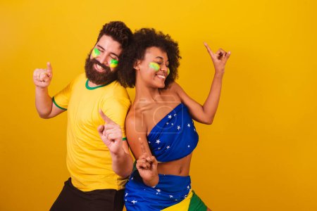 Téléchargez les photos : Couple de fans de football brésilien, habillé dans les couleurs du brésil, femme noire, homme caucasien. danse, s'amuser. - en image libre de droit