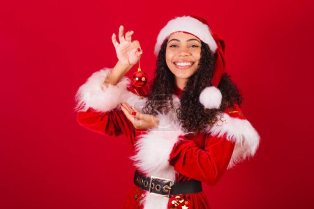Photo for Beautiful brazilian black woman, dressed as santa claus, christmas clothes. holding red ball, christmas ornament - Royalty Free Image