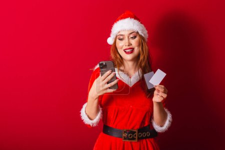 Téléchargez les photos : Belle femme rousse brésilienne, vêtue de vêtements de Noël, le Père Noël. tenant des cartes de crédit et smartphone. - en image libre de droit