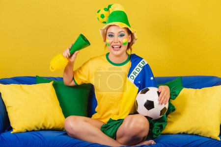 Photo for Caucasian redhead woman brazil soccer fan on couch with horn and ball cheering - Royalty Free Image