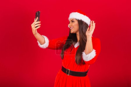 Photo for Beautiful Brazilian woman dressed in Christmas clothes, Santa Claus. holding smartphone video call. - Royalty Free Image