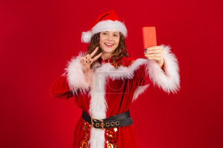 Photo for Brazilian woman, dressed in Santa Claus outfit, holding smartphone taking self portrait. - Royalty Free Image
