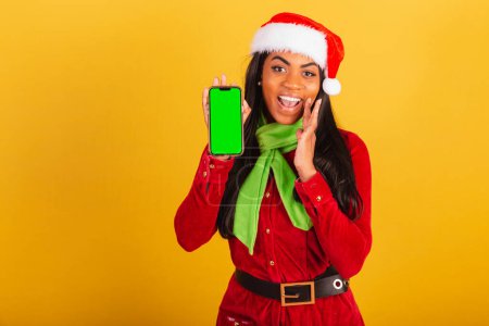 Photo for Beautiful black Brazilian woman, dressed in christmas clothes, santa claus, smartphone and showing green chroma screen. - Royalty Free Image