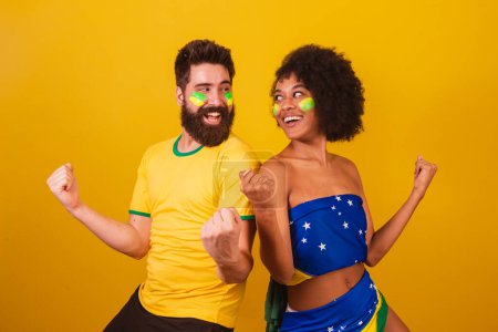 Téléchargez les photos : Couple de fans de football brésilien, habillé dans les couleurs du brésil, femme noire, homme caucasien. - en image libre de droit