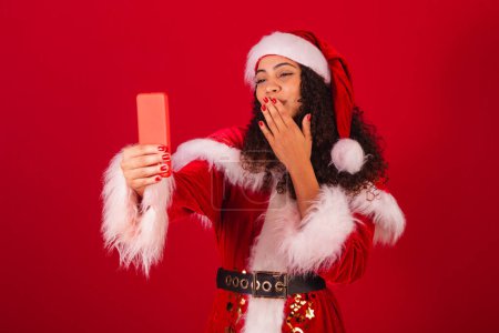 Photo for Beautiful brazilian black woman, dressed as santa claus, christmas clothes. in video call by smartphone. - Royalty Free Image