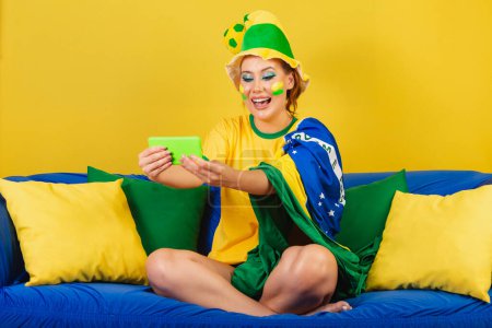 Photo for Caucasian woman, redhead, brazil soccer fan, brazilian, on couch watching game by smartphone - Royalty Free Image