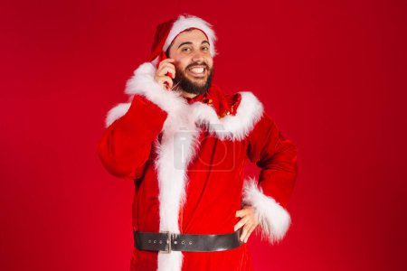Photo for Brazilian man dressed in santa claus clothes holding smartphone on voice call - Royalty Free Image