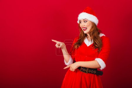Téléchargez les photos : Belle femme rousse brésilienne, vêtue de vêtements de Noël, le Père Noël. montrer quelque chose à gauche. - en image libre de droit