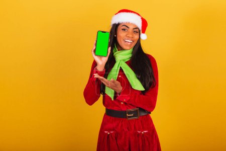 Téléchargez les photos : Belle femme brésilienne noire, vêtue de vêtements de Noël, Père Noël claus, smartphone et montrant écran chromé vert. - en image libre de droit