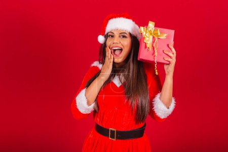Foto de Hermosa mujer brasileña vestida con ropa de Navidad, Santa Claus. sorprendido y pensando en rojo presente. - Imagen libre de derechos