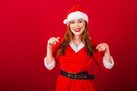 Photo for Beautiful Brazilian red-haired woman, dressed in Christmas clothes, Santa Claus. only here, pointing towards the center. - Royalty Free Image