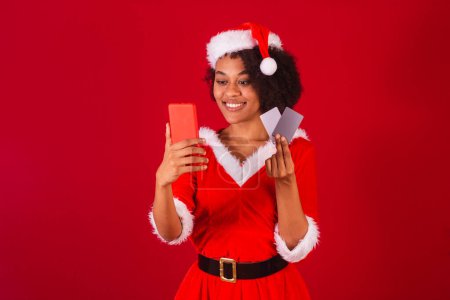 Téléchargez les photos : Belle femme brésilienne noire, habillée en Père Noël claus, maman claus, tenant smartphone et cartes de crédit - en image libre de droit