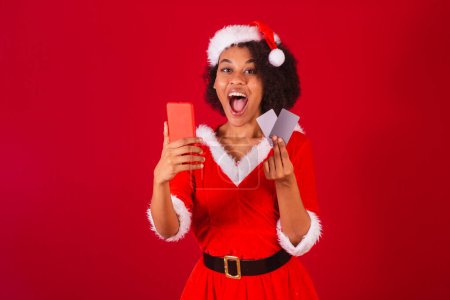 Téléchargez les photos : Belle femme brésilienne noire, habillée en Père Noël claus, maman claus, tenant smartphone et cartes de crédit - en image libre de droit
