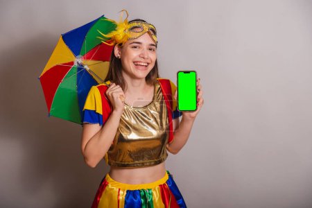 Foto de Hermosa brasileña, mujer caucásica, vistiendo frevo, ropa de carnaval, usando una máscara. sostener smartphone con pantalla verde en croma. - Imagen libre de derechos