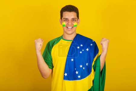 Foto de Joven aficionado al fútbol brasileño. vestido de verde, mano sobre pecho, celebrando con la bandera de Brasil. - Imagen libre de derechos