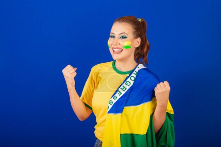 Foto de Mujer caucásica, pelirroja, aficionada al fútbol brasileño, brasileña, fondo azul, con bandera de Brasil, celebrando - Imagen libre de derechos