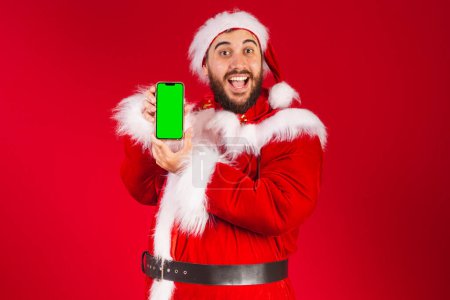 Photo for Brazilian man, dressed in santa claus clothes, holding smartphone with green chroma screen - Royalty Free Image