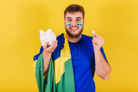 Téléchargez les photos : Homme caucasien brésilien, fan de football du Brésil, tenant tirelire, et pièce de monnaie - en image libre de droit