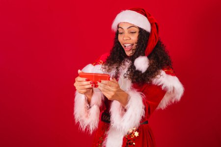 Foto de Hermosa mujer negra brasileña, vestida como santa claus, ropa de Navidad. celebrando con algo en el smartphone. - Imagen libre de derechos