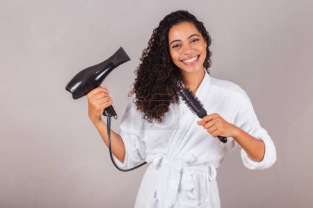 Photo for Beautiful Brazilian black woman, wearing bathrobe and towel. drying hair with dryer and brush, spa, beauty center, hair care. - Royalty Free Image