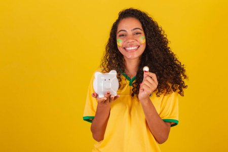 Photo for Young black Brazilian woman, soccer fan. holding piggy bank and coin. finance, economy, retirement. - Royalty Free Image