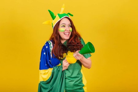 Foto de Mujer caucásica, pelirroja, fanática del fútbol brasileño, con cuerno, haciendo ruido, partido de fútbol. aplausos. - Imagen libre de derechos