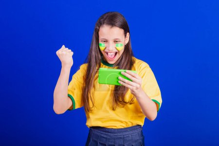 Photo for Young girl, soccer fan from Brazil. holding cellphone, watching game, cheering for Smartphone. applications. - Royalty Free Image