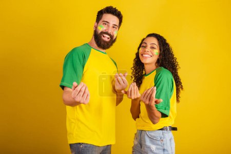 Téléchargez les photos : Couple de supporters brésiliens de football, vêtus aux couleurs de la nation, femme noire, homme caucasien. invitant avec les mains. - en image libre de droit