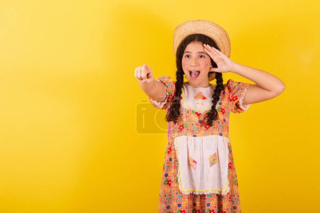 Téléchargez les photos : Fille portant des vêtements traditionnels orange pour la fête junina. Observation et pointage vers quelque chose de loin, incroyable. Promotion, publicité. - en image libre de droit